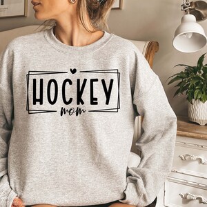 以下が含まれることがあります： 黒い枠の中に「HOCKEY mom」という文字が入ったグレーのトレーナー。文字の上に小さなハートマークがあります。