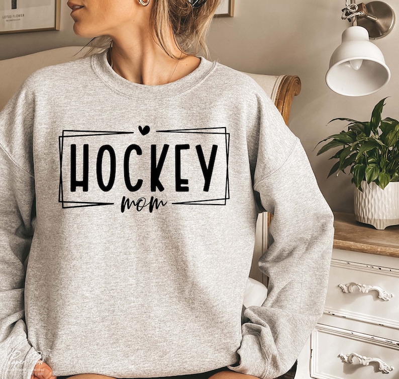 以下が含まれることがあります： 黒い枠の中に「HOCKEY mom」という文字が入ったグレーのトレーナー。文字の上に小さなハートマークがあります。