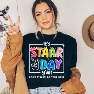 Può includere: Una maglietta nera con una scatola bianca che presenta il testo "It's STAAR DAY y'all DON'T STRESS! DO YOUR BEST" in lettere arcobaleno. Il testo è in un divertente font da cartone animato.