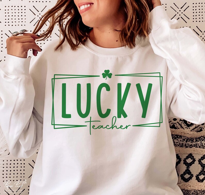 Puede incluir: Sudadera blanca con un tr&eacute;bol verde y el texto "LUCKY teacher" en un cuadro verde.