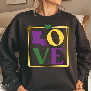 May include: Black sweatshirt with a graphic print of the word "LOVE" in purple, green, and yellow. The letter "O" is replaced with the outline of the state of Louisiana. A small green heart is above the graphic.