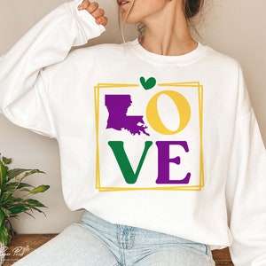 May include: White sweatshirt with a graphic design of the word "LOVE" in purple, green, and yellow. The "O" is replaced with the outline of the state of Louisiana in purple. A small green heart is above the graphic.