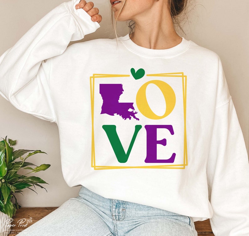 May include: White sweatshirt with a graphic design of the word "LOVE" in purple, green, and yellow. The "O" is replaced with the outline of the state of Louisiana in purple. A small green heart is above the graphic.