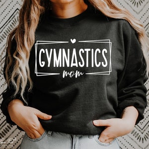 May include: Black sweatshirt with the words "GYMNASTICS mom" in white, inside a white rectangular frame. A small white heart is above the word "mom". The sweatshirt is a crew neck style.