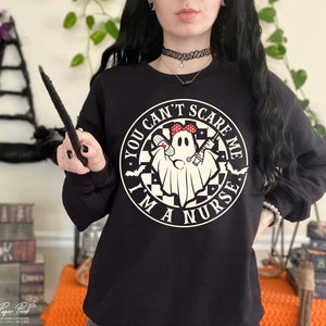 Puede incluir: Sudadera negra con un gr&aacute;fico circular blanco que dice "You Can't Scare Me I'm A Nurse" con un fantasma que lleva una bandana roja y sostiene una jeringa y un vendaje.