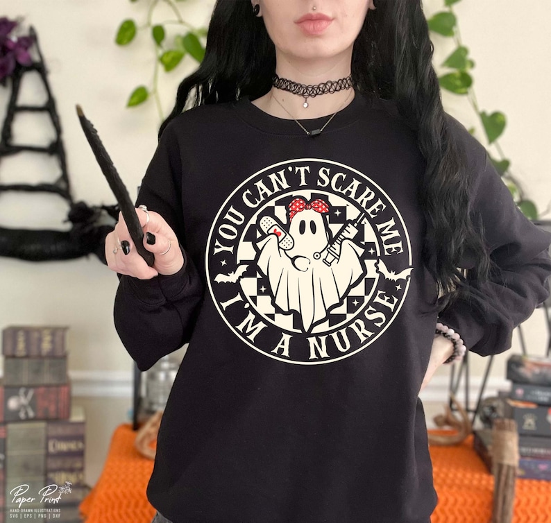 Puede incluir: Sudadera negra con un gr&aacute;fico circular blanco que dice "You Can't Scare Me I'm A Nurse" con un fantasma que lleva una bandana roja y sostiene una jeringa y un vendaje.