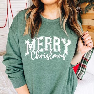 K&ouml;nnte beinhalten: Ein gr&uuml;ner Pullover mit dem Text "Merry Christmas" in wei&szlig;en Buchstaben. Der Text ist in einer Vintage-Schriftart im College-Stil.