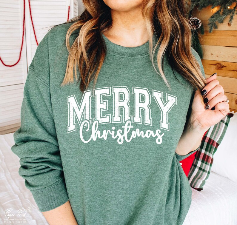 K&ouml;nnte beinhalten: Ein gr&uuml;ner Pullover mit dem Text "Merry Christmas" in wei&szlig;en Buchstaben. Der Text ist in einer Vintage-Schriftart im College-Stil.