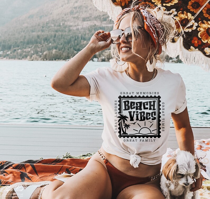 Puede incluir: Una mujer con una camiseta blanca con un gr&aacute;fico negro que dice "Beach Vibes" en un dise&ntilde;o de sello vintage. El gr&aacute;fico tambi&eacute;n incluye palmeras, un sol y el texto "Great Memories, Great Laughs, Great Times, Great Family".
