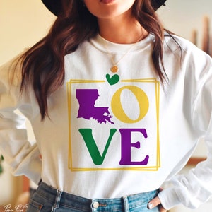 May include: White sweatshirt with a graphic design of the word "LOVE" in purple, green, and yellow. The "O" is replaced with the outline of the state of Louisiana in purple. A small green heart is above the graphic.