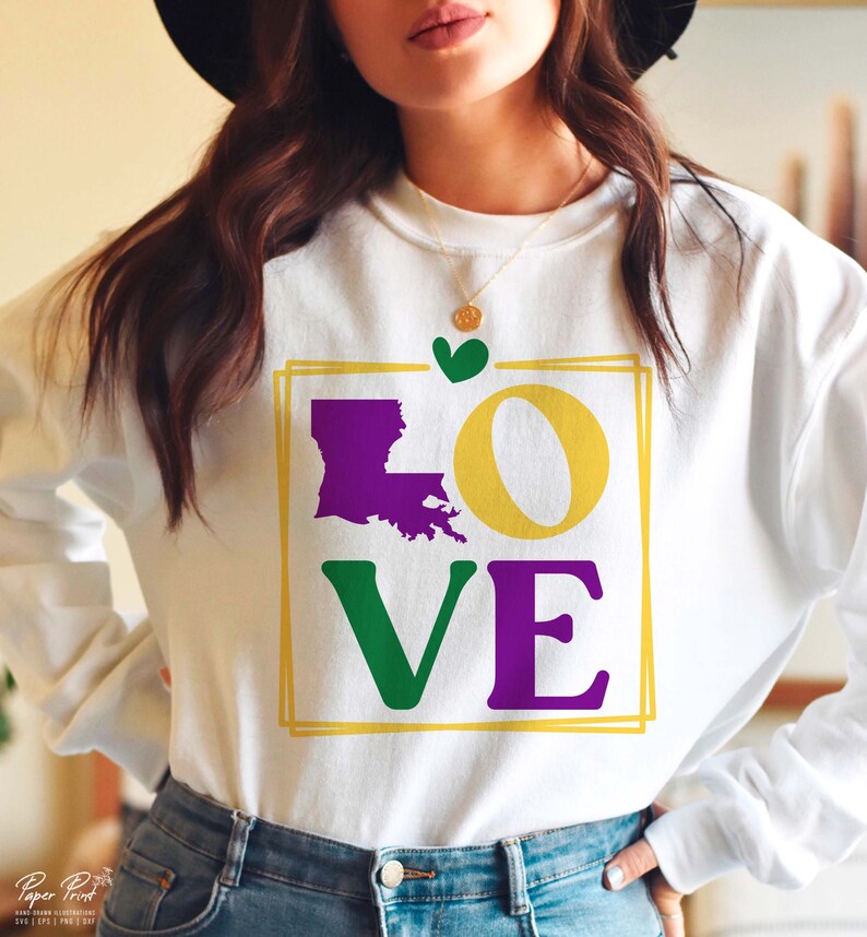 May include: White sweatshirt with a graphic design of the word "LOVE" in purple, green, and yellow. The "O" is replaced with the outline of the state of Louisiana in purple. A small green heart is above the graphic.