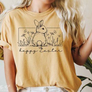 May include: A yellow t-shirt featuring a black line drawing of a bunny rabbit in a field of flowers. The text "happy Easter" is written below the bunny.