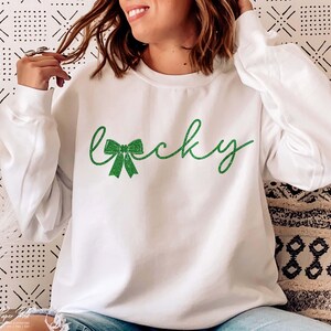 Puede incluir: Sudadera blanca con un dise&ntilde;o de purpurina verde que dice "lucky" con un lazo de purpurina verde.
