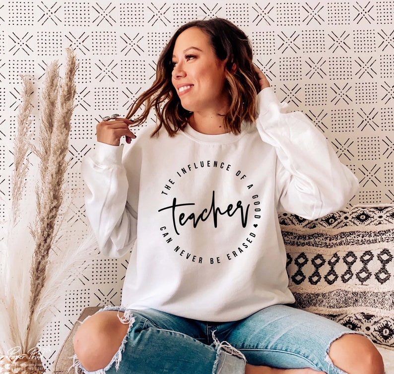 May include: White sweatshirt with a black circle graphic that says "The influence of a good teacher can never be erased".