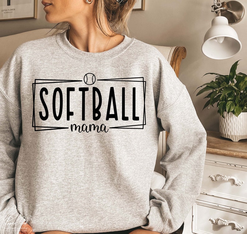 Pu&ograve; includere: Felpa grigio chiaro con la scritta nera "SOFTBALL mama" all'interno di un rettangolo nero, con una grafica di softball sopra. La felpa ha maniche lunghe e girocollo.