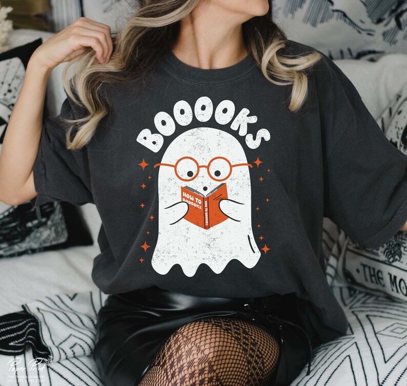 Pode incluir: Uma camiseta cinza escura com um fantasma branco usando &oacute;culos e lendo um livro que diz "How to Spook" com o texto "BOOooks" acima do fantasma.