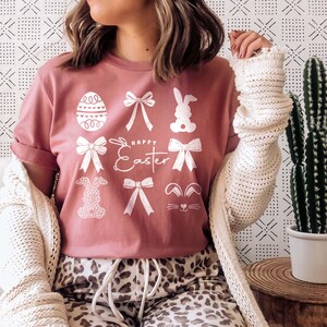 May include: A pink t-shirt with a white graphic design featuring bunnies, bows, and an Easter egg. The text "Happy Easter" is printed on the shirt.