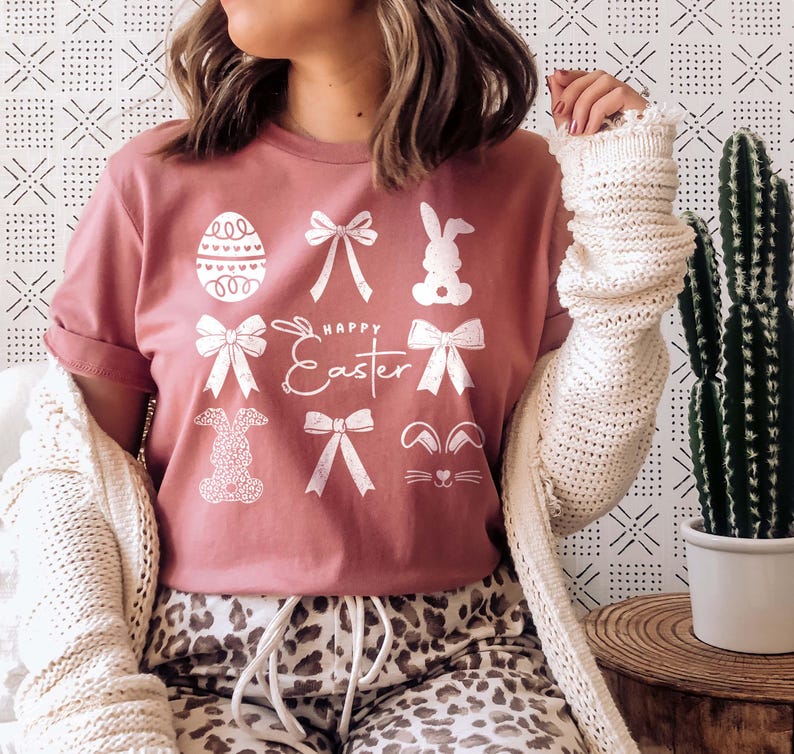 May include: A pink t-shirt with a white graphic design featuring bunnies, bows, and an Easter egg. The text "Happy Easter" is printed on the shirt.