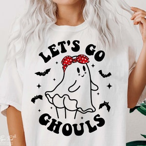 May include: White t-shirt with a black and white illustration of a ghost wearing a red and white bandana. The text "Let's go ghouls" is printed on the shirt.