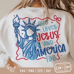 Puede incluir: Una camiseta blanca con un dise&ntilde;o gr&aacute;fico rojo, blanco y azul. El gr&aacute;fico presenta a una mujer vestida como la Estatua de la Libertad sosteniendo una antorcha. El texto "She Loves Jesus And America Too" est&aacute; escrito en letras rojas, blancas y azules.