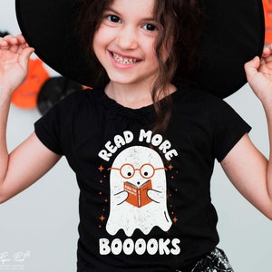 May include: A black t-shirt with a white ghost wearing glasses and reading a book that says "How to Scare" with the text "Read More Booooks" above the ghost.