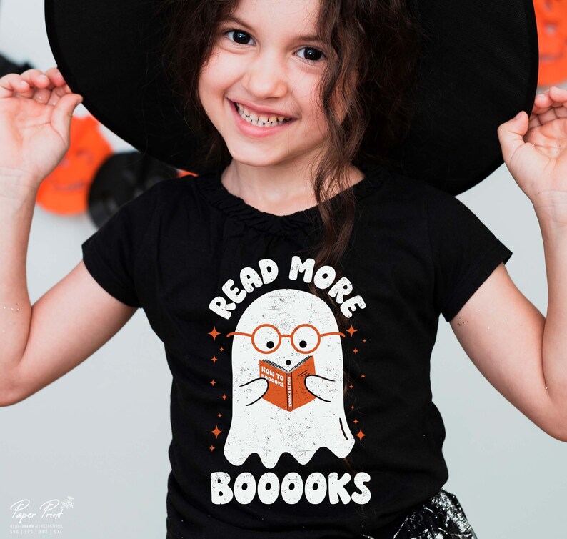 May include: A black t-shirt with a white ghost wearing glasses and reading a book that says "How to Scare" with the text "Read More Booooks" above the ghost.