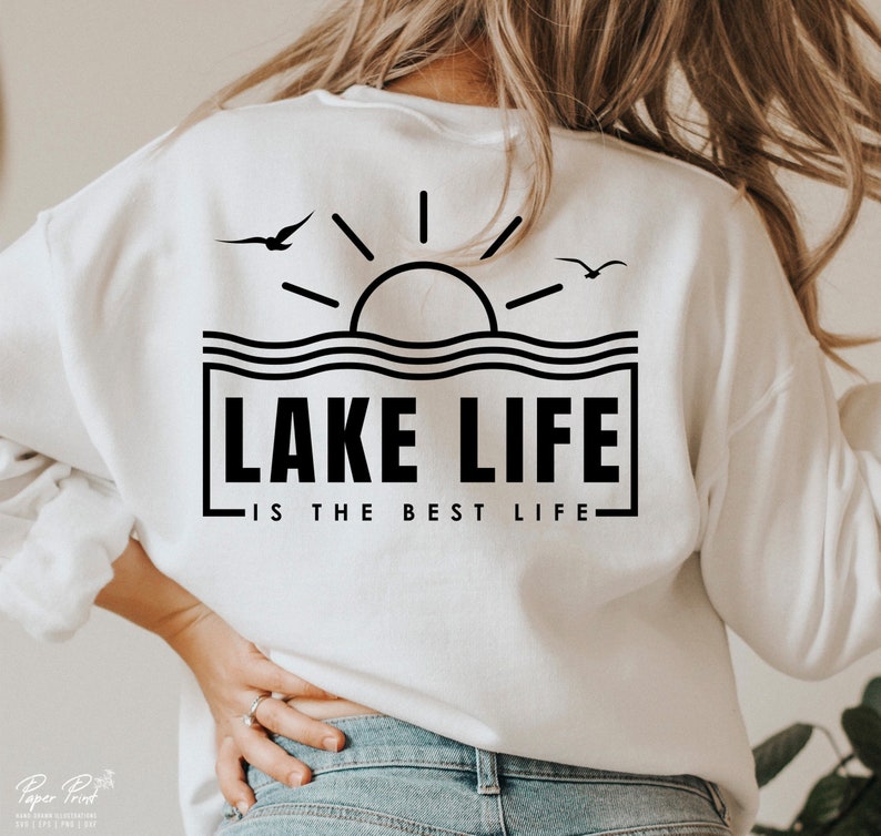 May include: White sweatshirt with a black graphic design on the back. The design features a sun, waves, and the text "LAKE LIFE IS THE BEST LIFE" in a rectangular frame, with two birds flying above the waves.