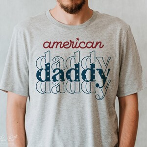 Puede incluir: Una camiseta gris con el texto "american daddy" impreso en ella. La palabra "daddy" se repite tres veces, con la "daddy" del medio con un patr&oacute;n de estrellas blancas sobre un fondo azul.
