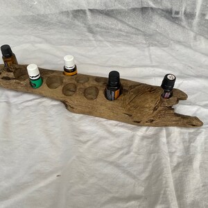 May include: A rustic wooden essential oil holder with 10 slots. The holder is made from driftwood and has a natural, weathered finish. It is filled with various essential oil bottles, including green, orange, and black.