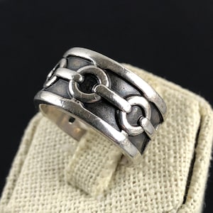 May include: A silver ring with a chain link design. The ring has a wide band with a series of interconnected chain links. The metal has a slightly oxidized appearance, giving it a vintage look. The ring is displayed on a neutral-colored surface.