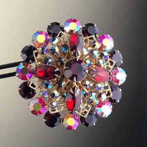 Op de afbeelding: Een grote, goudkleurige broche met een bloemmotief. De broche is versierd met rode, paarse en iriserende strasssteentjes.