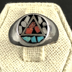 May include: A silver ring with a geometric design. The ring features a central triangle filled with red and white stones, surrounded by black, with turquoise stones below. The ring is set on a textured, off-white surface.