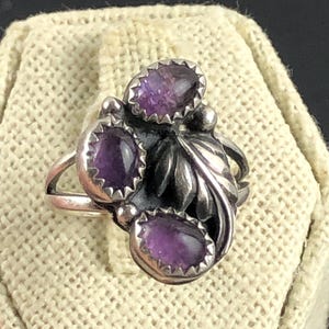 May include: A silver ring featuring three oval-shaped, deep purple amethyst stones. The ring has a floral design with a leaf and small silver beads. The ring is set on a textured, off-white surface.