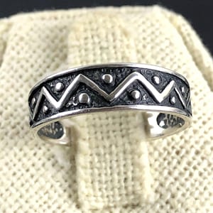 May include: A silver ring with a dark, textured band featuring a zigzag pattern and small circular accents. The ring is displayed on a cream-colored, textured surface, showcasing its intricate design and craftsmanship.
