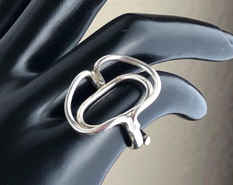 Puede incluir: Un anillo plateado con un diseño abstracto. El anillo presenta un diseño abierto y en bucle con un acabado liso y pulido. El anillo se muestra sobre una superficie negra.