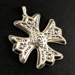 May include: A silver-colored pendant in the shape of a cross. The pendant has a textured surface with raised circular details and a loop at the top for a chain. The cross has four curved arms and a central floral design.