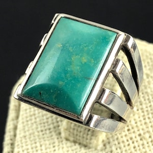 Op de afbeelding: Een zilveren ring met een rechthoekige turkoois steen. De turkoois heeft een levendige blauwgroene kleur met natuurlijke variaties. De ring heeft een geometrisch ontwerp met meerdere zilveren banden aan de zijkanten.