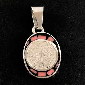 May include: Silver pendant with a black and pink enamel design. The pendant features a circular design with a sun-like pattern in the center. The pendant is oval shaped and has a bail for attaching to a chain.