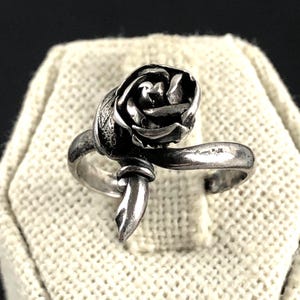 Puede incluir: Un anillo plateado con un diseño de rosa detallado. La rosa es el punto focal, con intrincados detalles de pétalos. La banda se curva alrededor del dedo, y el anillo tiene una estética vintage o antigua.