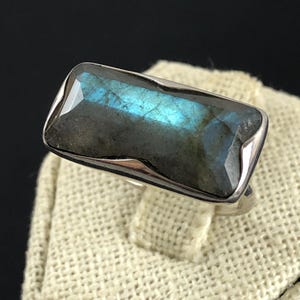 Puede incluir: Anillo de plata con una piedra labradorita rectangular. La piedra tiene una base gris oscuro con destellos iridiscentes azules y verdes. El anillo está sobre una superficie beige claro sobre un fondo oscuro.