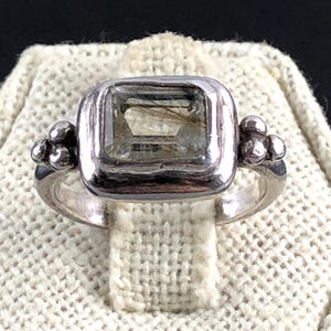May include: A silver ring with a square, light green gemstone. The ring has a rounded band and small, round embellishments on each side of the gemstone. The ring is displayed on a white, textured surface.