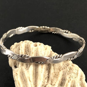 Op de afbeelding: Een zilverkleurige armband met een gedraaid touwontwerp. De armband ligt op een stuk lichtgekleurd koraal, met een donkere achtergrond. De armband heeft een eenvoudig, elegant ontwerp.