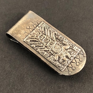 May include: A silver-toned money clip with an embossed rectangular design featuring an intricate pattern. The clip has a curved shape with decorative elements along the edges, and a spring-loaded mechanism.