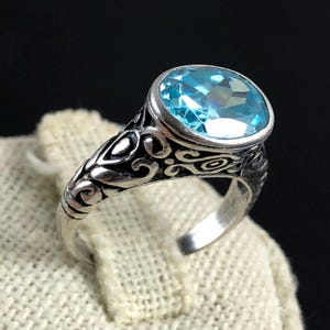May include: A silver ring with an oval, light blue gemstone. The ring has an ornate, swirling design on the band and around the gemstone setting. The gemstone is faceted, catching the light.