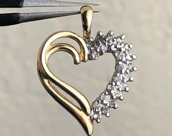 May include: A gold heart pendant with a diamond-studded edge. The pendant is held by tweezers, showcasing its intricate design. The heart shape is open, with a smooth, polished gold finish on one side and sparkling diamonds on the other.