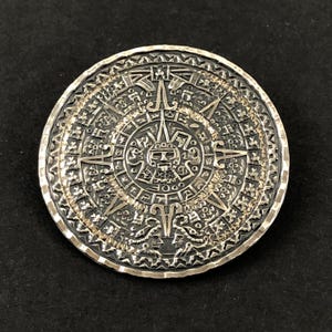 May include: A circular silver-toned metal medallion featuring an intricate, embossed Aztec calendar design. The center displays a stylized face surrounded by concentric rings of detailed geometric patterns and symbols with a dark, oxidized finish.