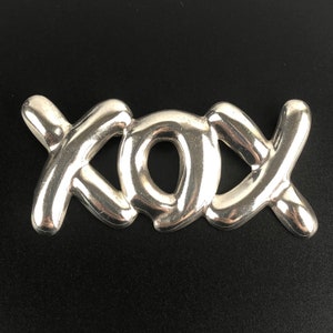 Op de afbeelding: Een zilveren bedel in de vorm van de letters "XOX" die in elkaar zijn gevlochten. De bedel is glad en glanzend.