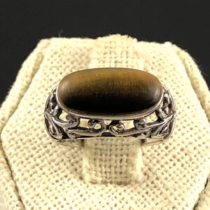 May include: A silver ring with an oval, brown tiger's eye stone. The ring has an ornate, openwork design around the stone. The ring is displayed on a cream-colored, textured surface.