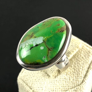 Può includere: Anello in argento con una grande pietra verde ovale con inclusioni marroni e bianche. La pietra è incastonata in una lunetta d'argento. L'anello è esposto su una superficie neutra. Design semplice ed elegante.