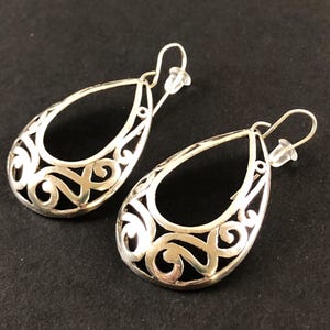 May include: A pair of silver teardrop earrings with an open filigree design. The earrings have a hook closure and a clear plastic backing. The intricate scrollwork pattern is visible against a dark background.